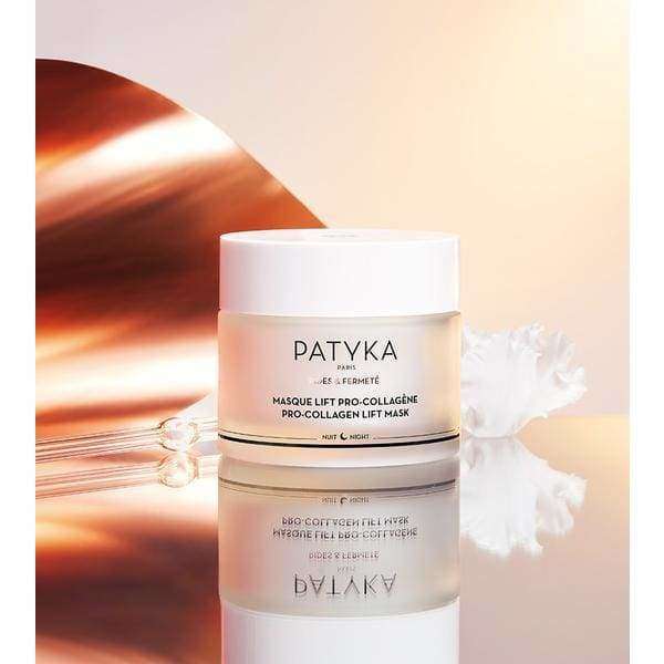Patyka Pro-Collagen Lift Mask Kasvonaamio; kiiltäväpintainen lieriömäinen purkki tekstillä "PATYKA Pro-Collagen Lift Mask" mustin groteskikirjaimin, seisoo peilipinnalla; purkki valkoinen kultaisella kannella; vieressä sileä valkoinen koristeellinen simpukankuoren muotoinen esine; tausta pehmeän vaaleanpunainen ja kultainen valaistus, ei ihmisiä tai käsiä; asetelmakuva.