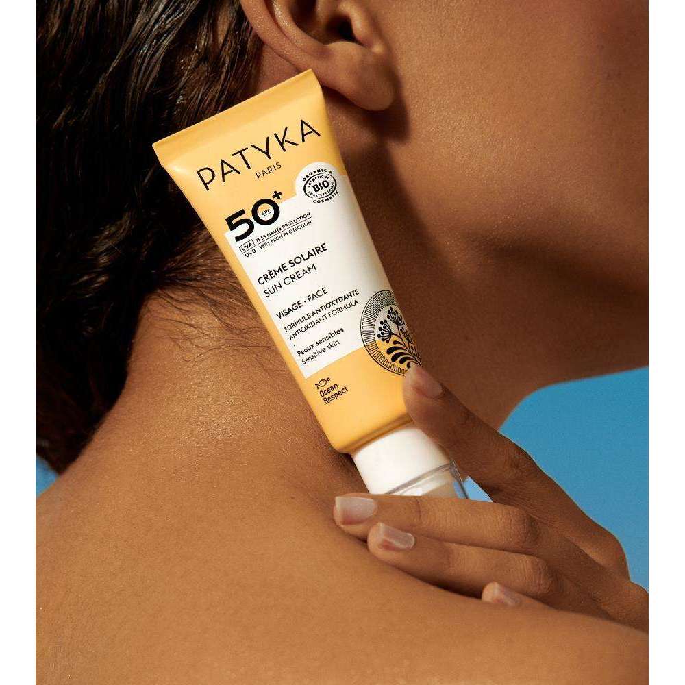 Patyka Face Sun Cream SPF50+ Aurinkovoide kasvoille; henkilö pitelee keltaista Patyka Paris SPF 50+ -aurinkovoidetuubia leukansa ja olkapäänsä lähellä sinistä taustaa vasten; tuubi pehmeää muovia mattapinnalla ja valkoinen läppäkorkki; etuetiketissä mustaa groteskitekstiä brändille ja lyhyelle tuotekuvaukselle sekä pieniä sertifiointilogoja; kuvassa vain henkilön käsi, osa leukaa ja kaulaa näkyvissä.