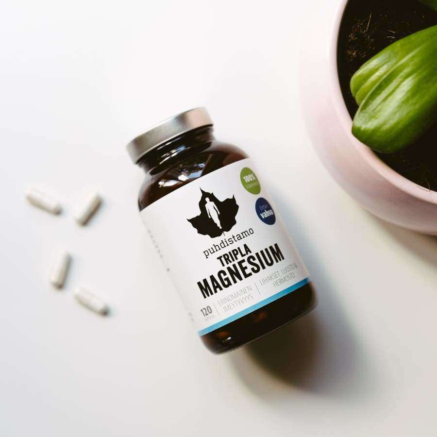 Puhdistamo Tripla Magnesium -pullo kyljellään, etiketissä teksti "Triple Magnesium" ja vihreä lehtilogo; kolme valkoista kapselia hajallaan vierellä; lisäksi pieni, mattapintainen vaaleanpunainen ruukku vihreälehtisen kasvin kanssa; kaikki aseteltu valkoiselle pinnalle, kuvattu ylhäältä tasaisessa valaistuksessa.