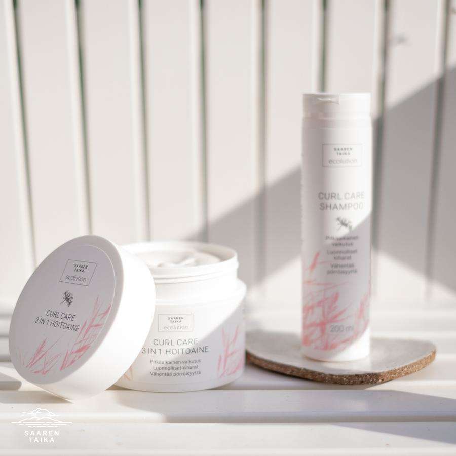 Saaren Taika Ecolution Hoitoaine Curl Care; kaksi hiustenhoitotuotetta auringonvalossa, avattu purkki paljastaa Curl Care 3 in 1 -hoitoaineen kermaisen sisällön ja korkea shampoopullo vierellä, molemmissa valkoiset ja vaaleanpunaiset etiketit; purkki matala ja leveä mattapintaista muovia, pullo kapea ja kiiltäväpintainen; moderni groteskikirjasin, tuotenimet luettavissa pastellitaustalla; ei ihmisiä kuvassa.