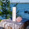 Saaren Taika Ecolution Shampoo Curl Care; pullo pumppuannostelijalla taitetulla pyyhkeellä karkeasti sahatun kannon päällä; pullo mattapintainen ja sylinterinmuotoinen, tekstinä "Curl Care Shampoo" lihavoidulla groteskikirjasimella; vieressä vaaleanpunainen kukka; taustalla tyyni järvi ja kirkas sininen taivas; ei ihmisiä tai käsiä kuvassa.