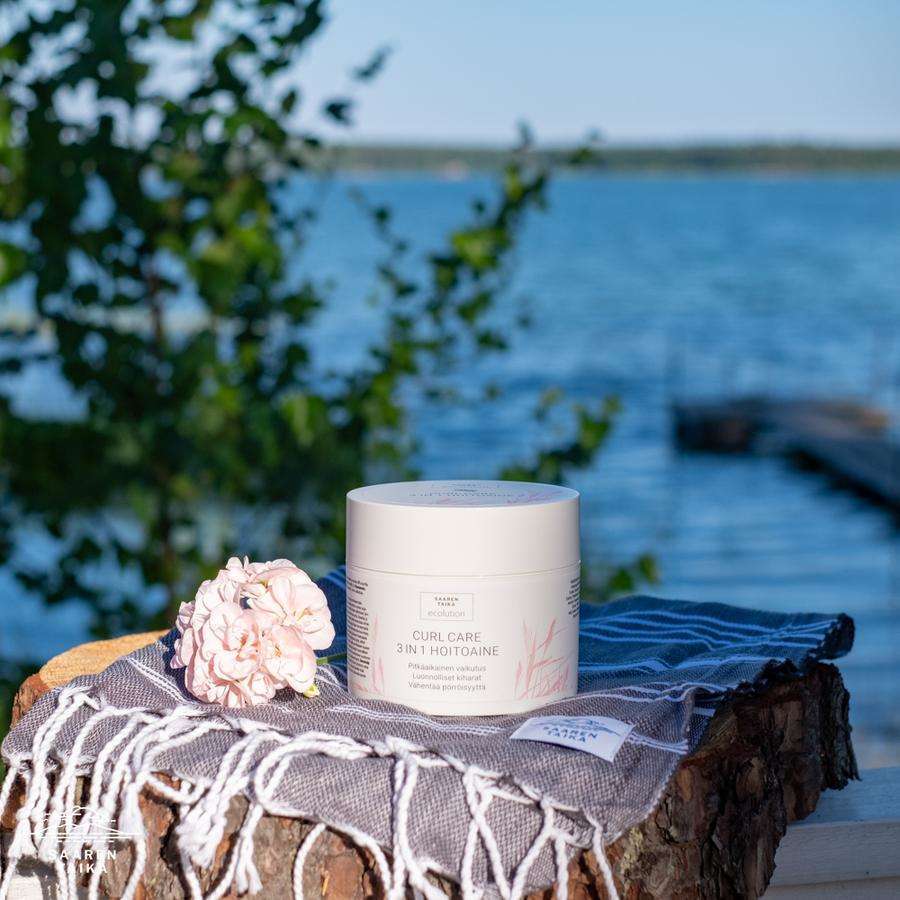 Saaren Taika Ecolution Hoitoaine Curl Care; purkki hapsureunaisella taitetulla pyyhkeellä pinottujen halkojen päällä, vieressä vaaleanpunainen kukka; purkki sylinterinmuotoinen kirkasta tai huurteista muovia yksivärisellä kierrekannella, mattapintainen; etiketissä yksinkertainen groteskiteksti; taustalla tyyni järvi ja puita, ei ihmisiä kuvassa.