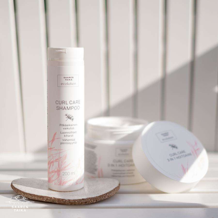 Saaren Taika Ecolution Shampoo Curl Care; kolme valkoista pulloa aseteltuna valkoiselle alustalle, yksi pystyssä ja kaksi kyljellään; kaikki mattapintaisia ja sylinterinmuotoisia; siniset groteskitekstit näyttävät tuotenimen kussakin pullossa; luonnollinen valaistus, ei ihmisiä kuvassa, neutraali tausta.
