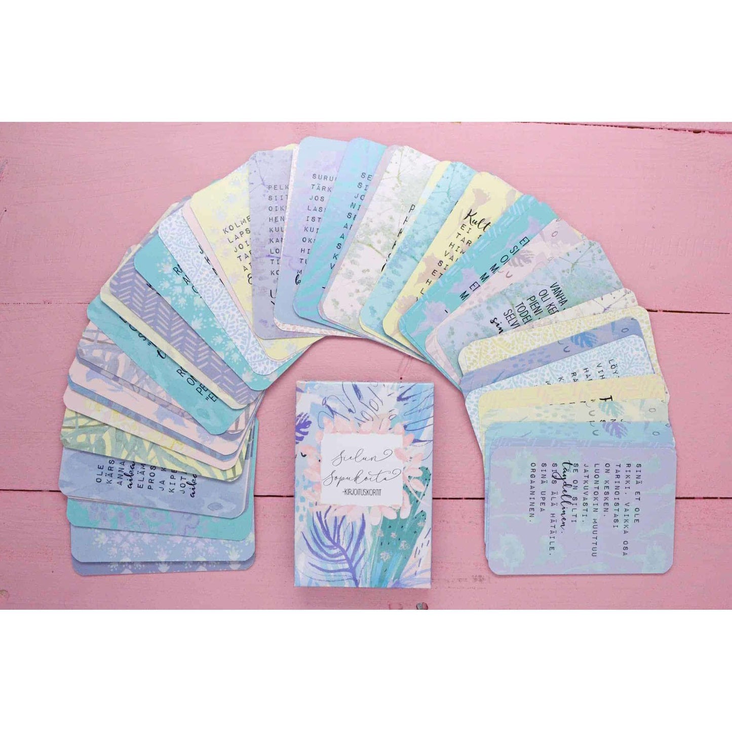 A set of colorful, pastel affirmation cards is arranged in a semicircle on a pink wooden surface, with one uplifting card deck box in the center. The cards have smooth matte finishes and rounded corners, displaying assorted soft colors including blue, green, yellow, and pink. The box matches the color scheme and sits upright. Typography varies by card but includes modern sans-serif fonts in white or dark hues. No people or hands are visible in the shot. Cards include legible phrases such as “You are