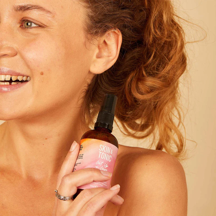 Skin & Tonic Jelly Cleanser puhdistusgeeli; kiharatukkainen nainen hymyilee ja pitää ruskeaa, mattapintaista ja lieriön muotoista pulloa kasvojensa lähellä, kyljessä valkoinen groteskiteksti "SKIN & TONIC"; beige tausta, kuvassa ylävartalo, kasvot ja toinen käsi.