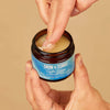 Skin & Tonic Calm Clean puhdistusbalmi; käsi ottaa balmia lieriön muotoisesta, mattapintaisesta ja sinisestä purkista, valkoisessa etiketissä lihavoitu musta-sininen groteskiteksti "Skin & Tonic Calm Clean Cleansing Balm"; beige tausta.