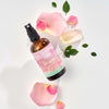 Skin & Tonic Be Soothed ruususuihke; ruskea suihkepullo valkoisella taustalla, tekstinä "Skin & Tonic Be Botanical Body & Room Mist"; musta, kiiltäväpintainen muovinen suihkeosa; ympärillä vaaleanpunaisia ruusunlehtiä ja tuoreita vihreitä lehtiä.