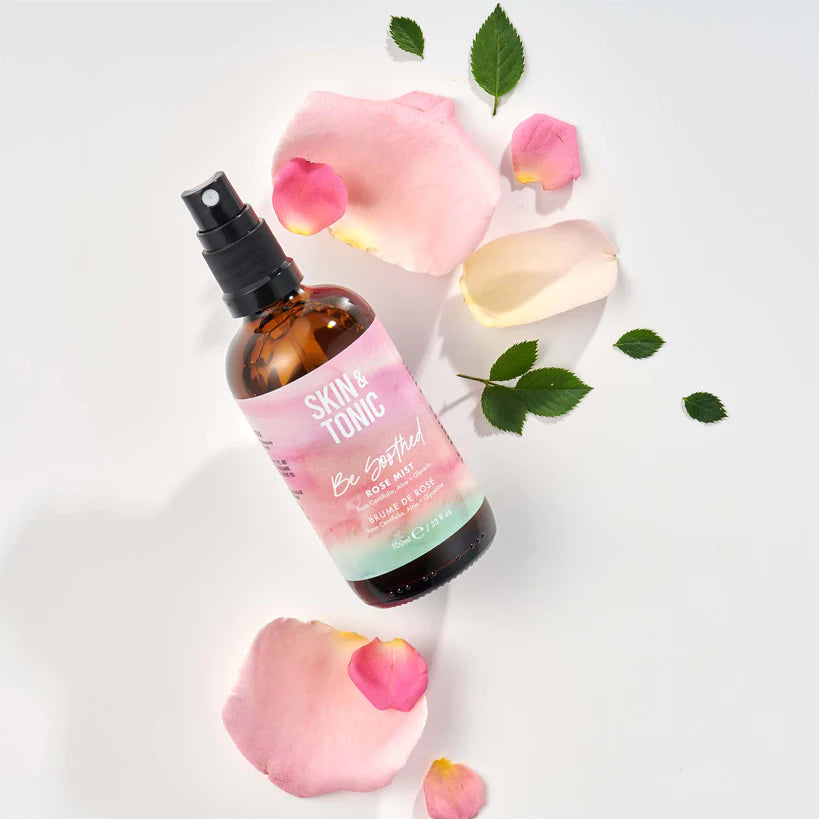 Skin & Tonic Be Soothed ruususuihke; ruskea suihkepullo valkoisella taustalla, tekstinä "Skin & Tonic Be Botanical Body & Room Mist"; musta, kiiltäväpintainen muovinen suihkeosa; ympärillä vaaleanpunaisia ruusunlehtiä ja tuoreita vihreitä lehtiä.