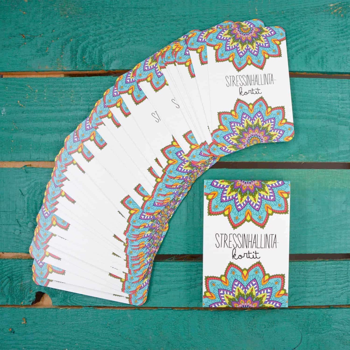 Hidasta Elämää Stressinhallintakortit; pakka värikkäitä tietoisuustaitokortteja monimutkaisin mandala-kuvioin levitettynä turkoosille puupinnalle; kortit suorakulmaisia mattapinnalla; päällimmäisessä kortissa lukee lihavoidulla groteskitekstillä "STRESSINHALLINTA kortit"; yksi lisäkortti lepää pääryhmän alla erillään viuhkasta; ei ihmisiä tai käsiä kohtauksessa.