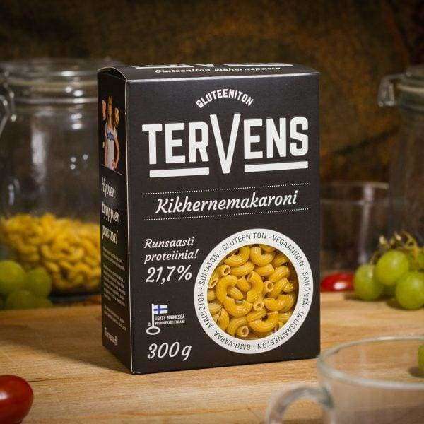 Tervens Kikhernemakaroni -pakkaus puisella pöydällä; pääosin keltainen pakkaus, jossa lihavoitua sinistä tekstausvapaata tekstiä sekä selkeät merkinnät korkeasta proteiinipitoisuudesta ja gluteenittomuudesta; ympärillä kirkkaita lasipurkkeja, vihreitä viinirypäleterttuja, yksittäinen punainen kirsikkatomaatti sekä läpinäkyvä lasikuppi; ei ihmisiä tai käsiä näkyvissä.
