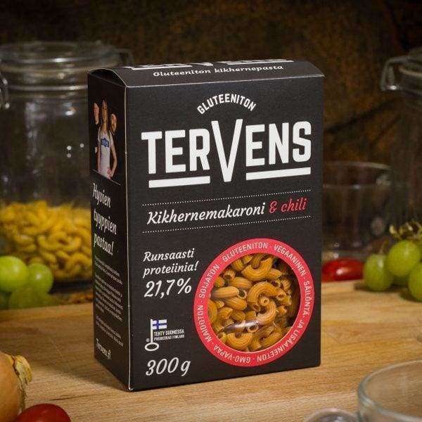 Tervens Kikhernemakaroni & Chili -pakkaus puisella pöydällä; ympärillä pastalla täytettyjä lasipurkkeja, vihreitä viinirypäleitä ja punaisia tomaatteja; pakkauksessa tekstausvapaata tekstiä, mattapintainen viimeistely sekä luettavissa teksti "gluten-free" ja "high protein"; ei ihmisiä kuvassa.