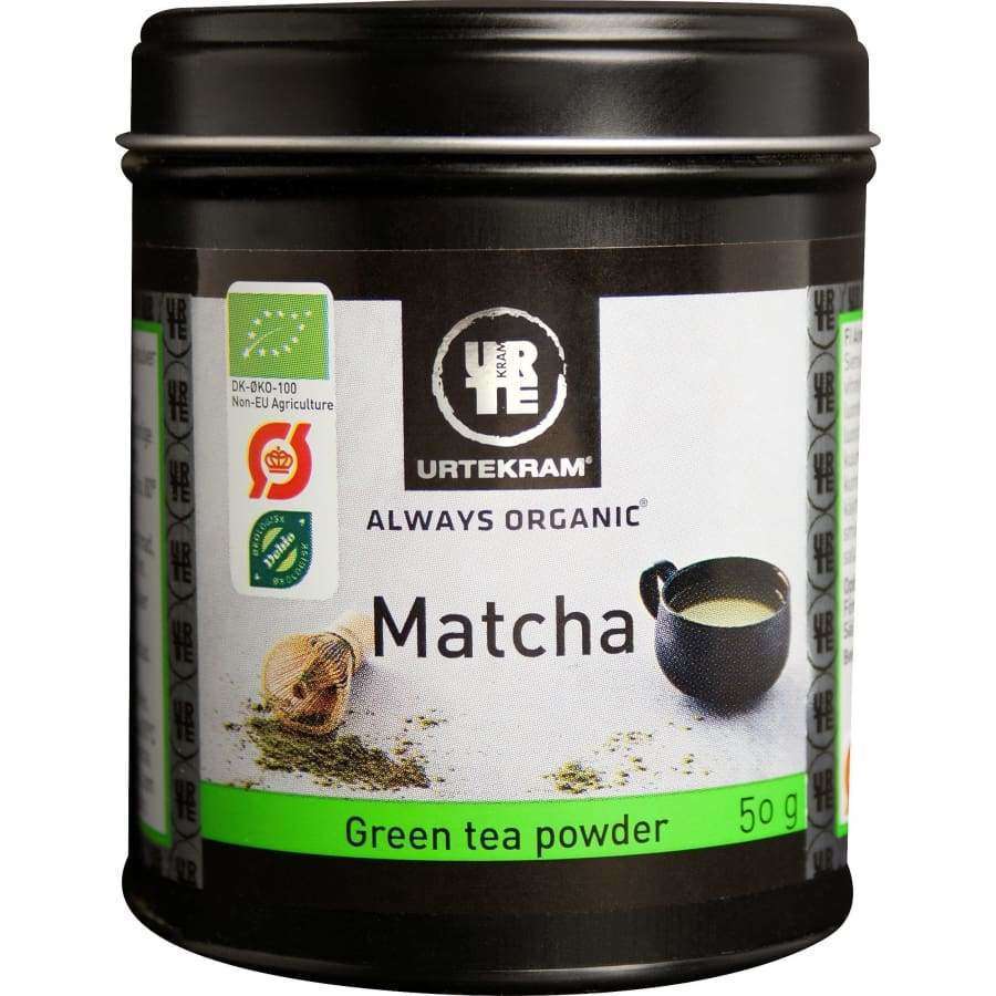 Urtekram Luomu Matcha-tee -metallipurkki, 50 g; musta, sylinterimäinen ja mattapintainen purkki sopivalla kannella; etiketissä kuva vihreää jauhetta sisältävästä kulhosta ja bambuvispilästä sekä vihreitä ja valkoisia värielementtejä, mukaan lukien EU:n luomusertifiointimerkki; lihavoitua valkoista tekstausvapaata tekstiä; kuvattu valkoista studiotaustaa vasten, ei ihmisiä tai käsiä näkyvissä.